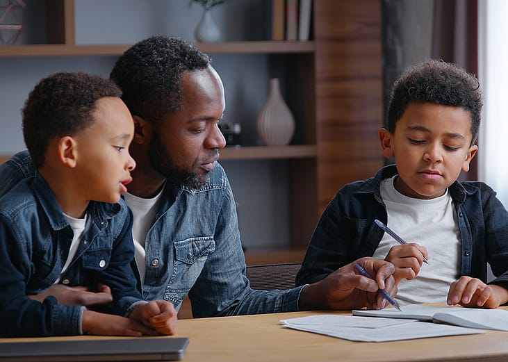 A parent studying with his young children, while helping his other son with a school writing assignment.
