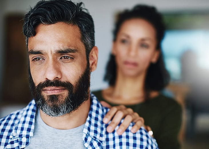 A man looking contemplative while a woman places a hand on his shoulder in a moment of emotional support.