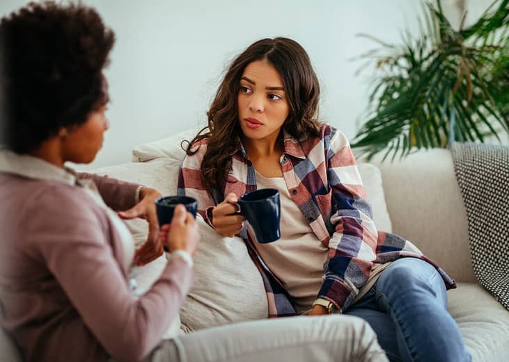 Two female friends having a discussion at home while drinking coffee.