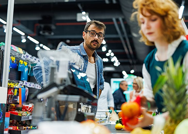 A male customer wearing glasses and denim shirt watches the cashier scanning groceries at a checkout.