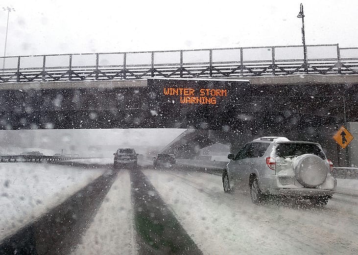 Windshield POV of a car passing under a bridge with an electronic 'Winter Storm Warning' sign.