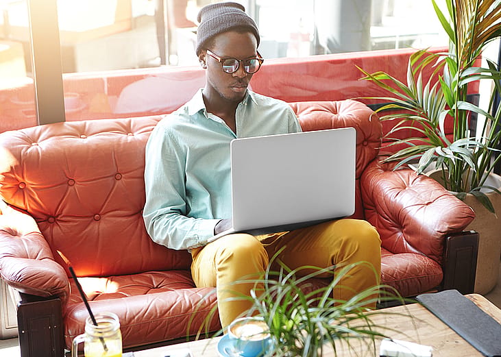 A fashionable young black freelancer working remotely on a laptop while sitting on a red leather sofa.