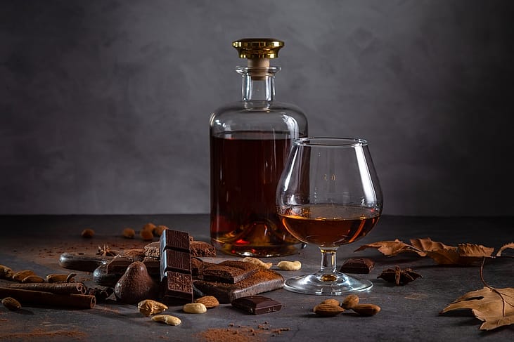 A glass of cognac, cognac in a bottle and pieces of chocolate on a dark background.