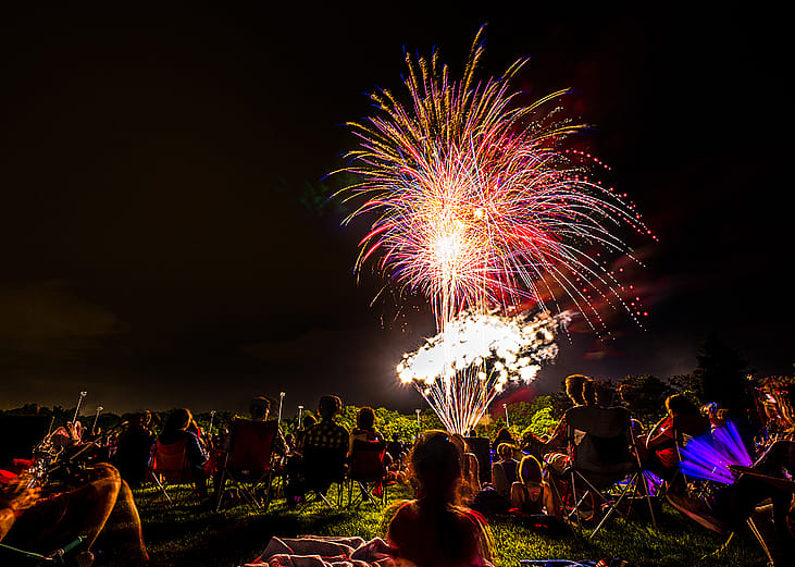 Crowds of people sit in lawn chairs and blankets to enjoy the colorful fourth of July independence day celebratory fireworks exploding in the black night sky.