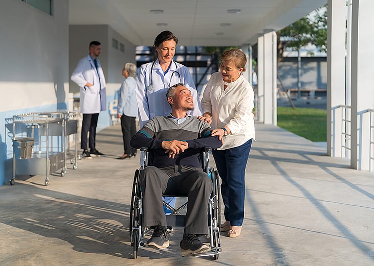 A female doctor assisting her patient in pushing his wheelchair along with a companion.