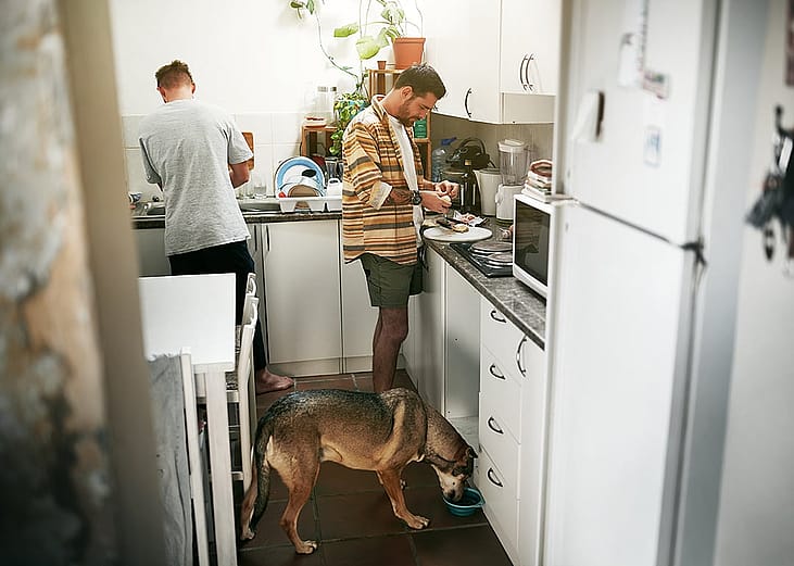 A young gay couple cooking together in their kitchen and feeding their dog.