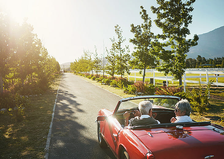An elderly couple driving in a convertible car for a road trip on a summer day.