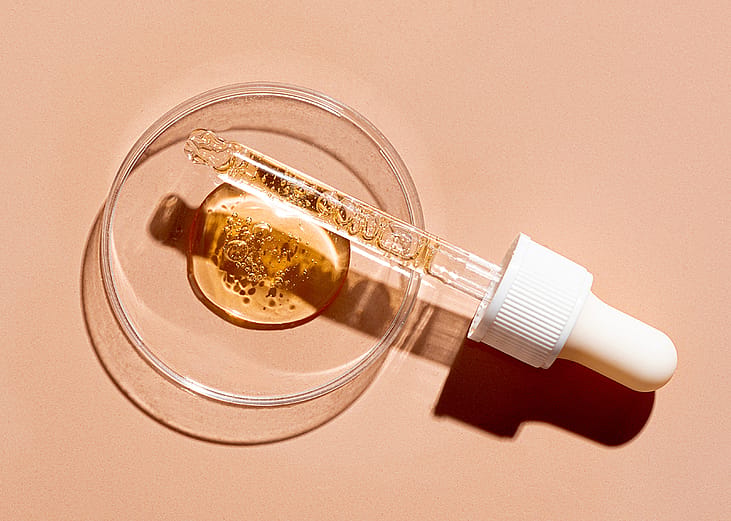 A dropper pipette and a petri dish with drops of oil or retinol gel on a beige nude background.