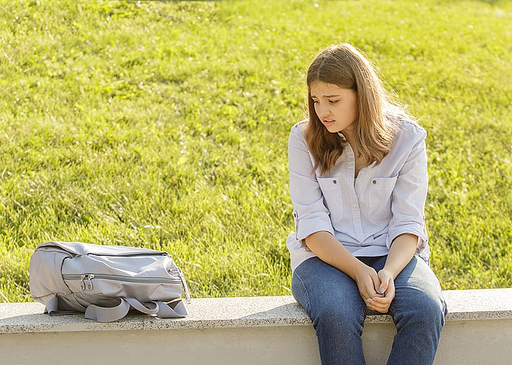 An upset teenage girl sitting on a curb and staring at her backpack for school.