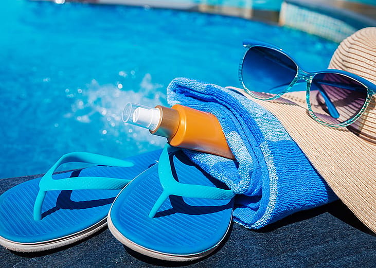 Slippers, sunscreen, body towel, straw hat, and sunglasses seen beside a swimming pool.