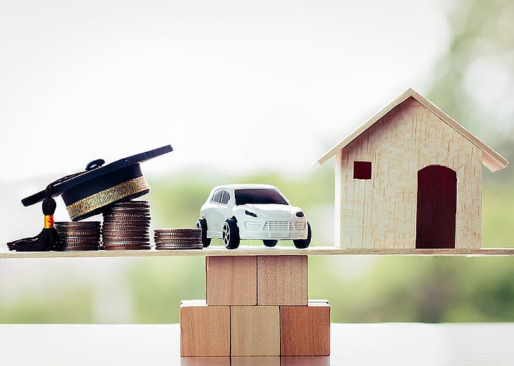 Miniatures of a graduation cap on top of coin stacks, a car, and a house above a wooden block, symbolizing the concept of success in university education.
