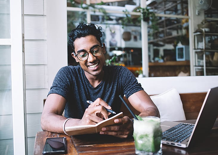 A happy young man taking notes while studying at a cafe.