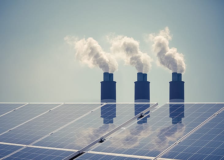 Photovoltaic solar panels with industrial smokestacks emitting smoke and steam in the background.