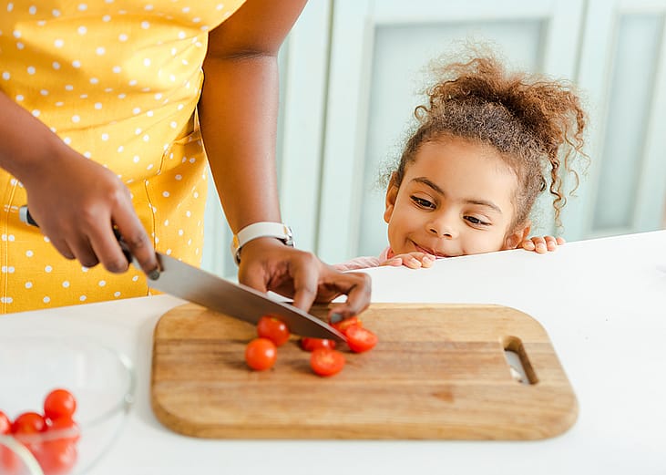 A little child watching her mother slice cherry tomatoes on a chopping board in the kitchen.