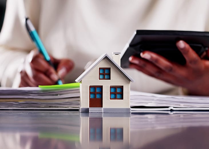 A small figure of a home with a real estate professional calculating finances in the background.