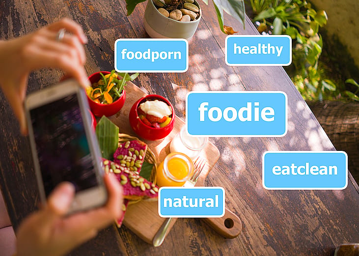 Person taking a photo of their food illustrated with common social media hashtags.