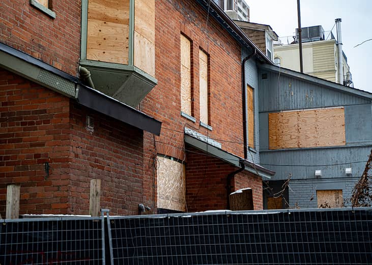 Abandoned brick and wooden houses with boarded windows enclosed by a construction fence.