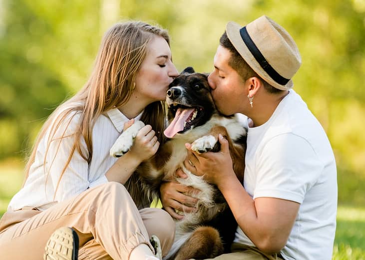 A young couple kissing their pet dog outdoors in a park.