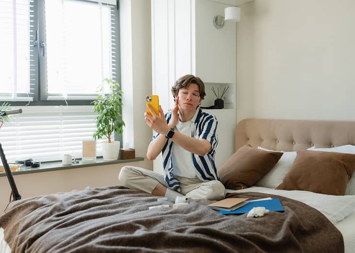 A young adult checking his face through his smartphone's front camera in a bedroom.