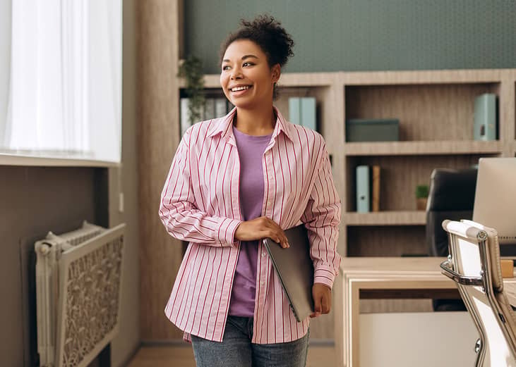 A happy businesswoman in an office walking with her laptop.