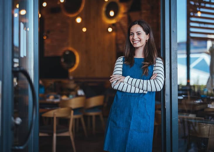 A female coffee shop owner happily stands at the entrance of her shop to welcome customers.