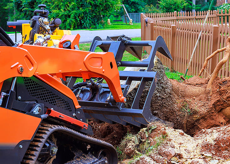 A compact track loader extracting large tree stump from a backyard in a suburban neighborhood.