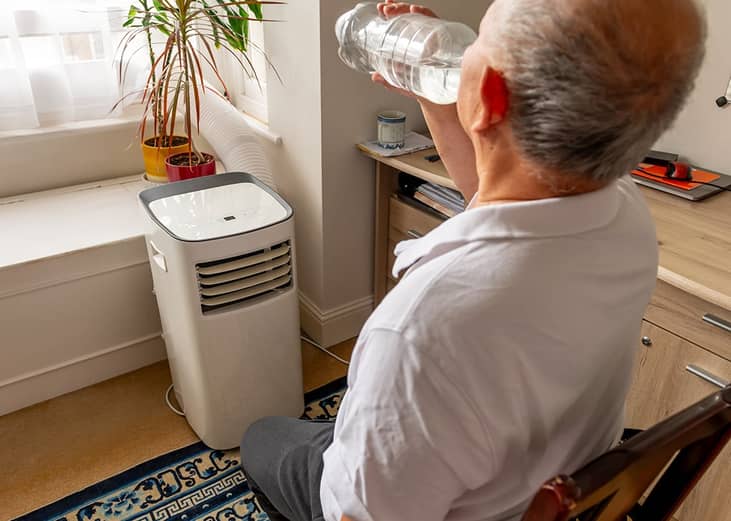 An elderly man drinking water and cooling himself in front of an AC unit during summer.