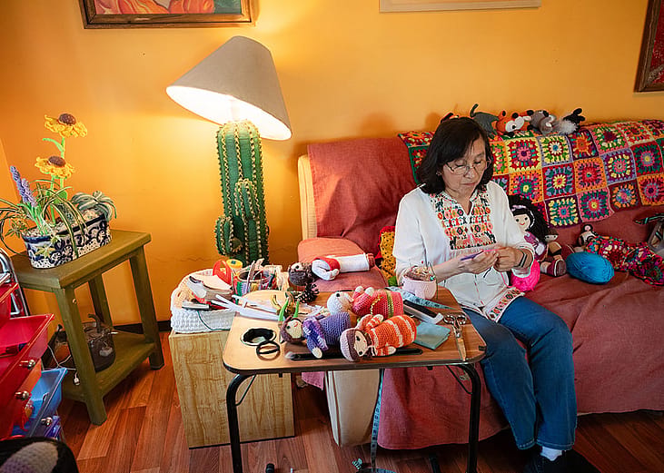 An elderly person crocheting a doll on a sofa, while surrounded by her other handmade creations at home.