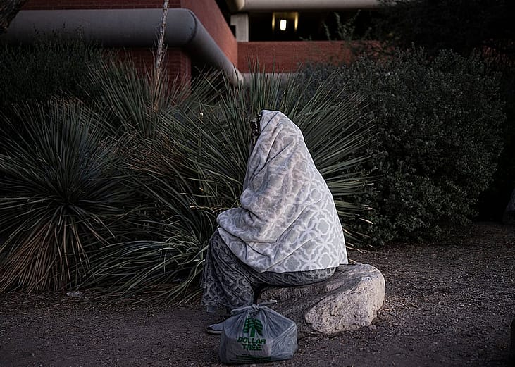 An unhoused person wrapped in blanket and sitting on a rock outdoors in Tucson, Arizona.