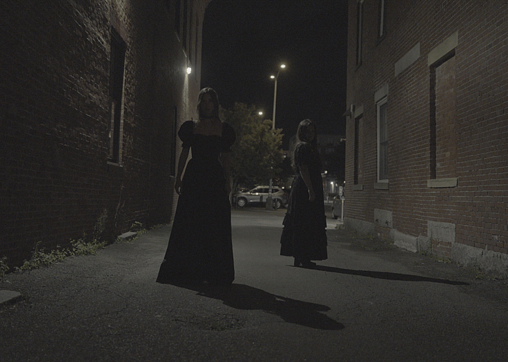 Two figures in black dresses stand in a dark brick alleyway at night.