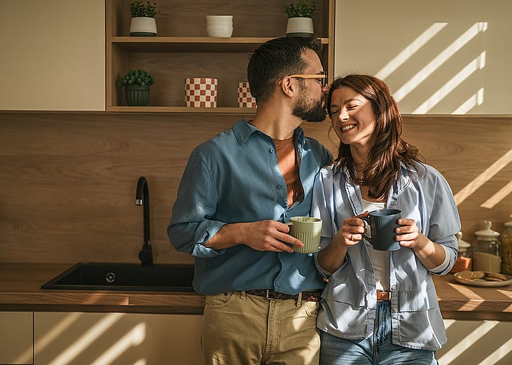 A happy couple hugging and drinking coffee together in their sun-lit kitchen.