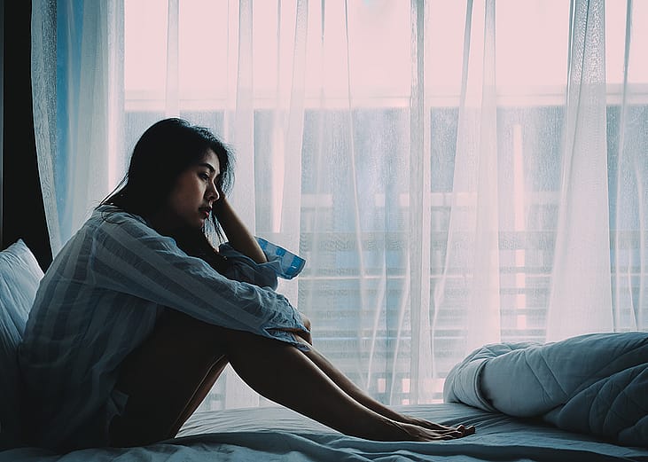 A young adult sitting on a bed and looking pensive near a window in her room.
