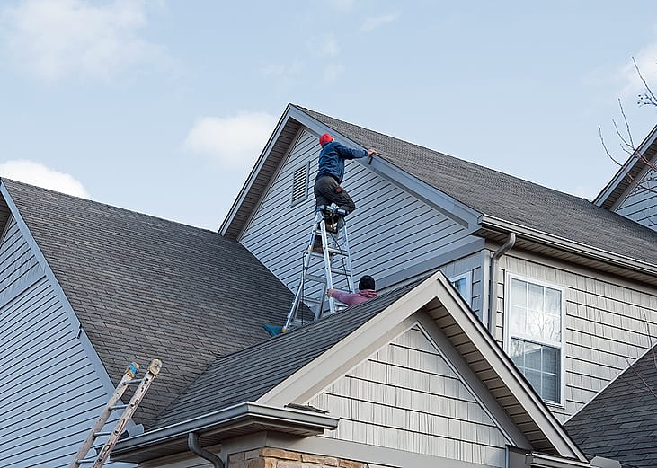 Professional roofers repairing damaged siding trim along multiple roof lines after storm damage.