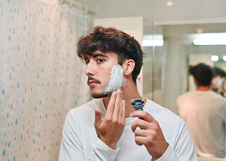A young man shaving his beard in front of a mirror.