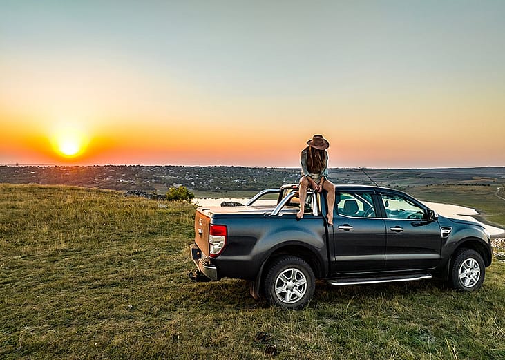 A person sitting on the roof of a pickup truck parked on a grassy hill during sunset. 