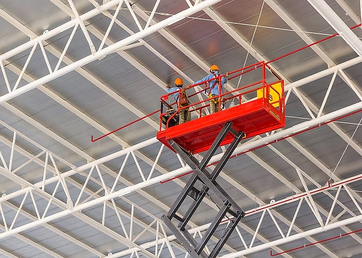 Construction workers doing a roof work in an elevated scissor lift platform with hydraulic system at a factory.