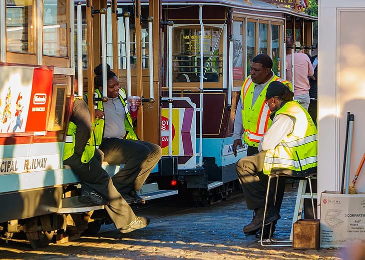 Employees in safety vests are talking and resting on a sunny day beside classic cable cars of the San Francisco Municipal Railway.