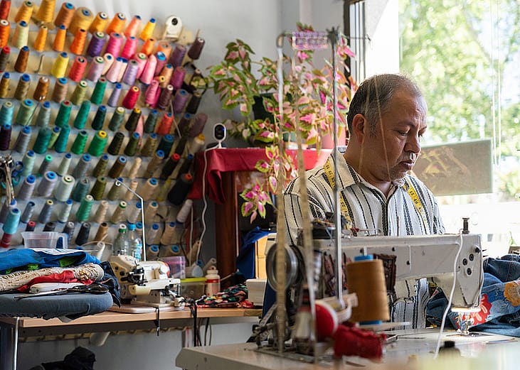 A tailor working with a sewing machine with colorful threads and fabric in the background at his shop.