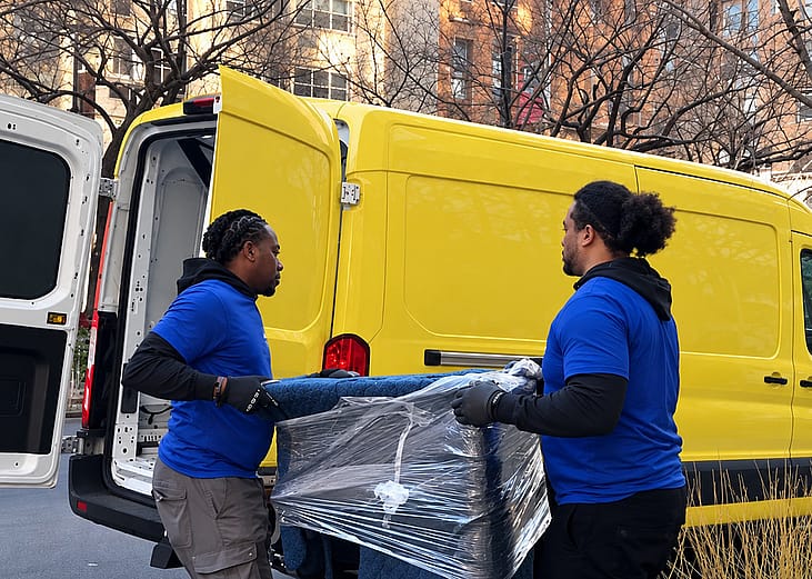 Two moving company workers load a piece of plastic-wrapped furniture for transport.