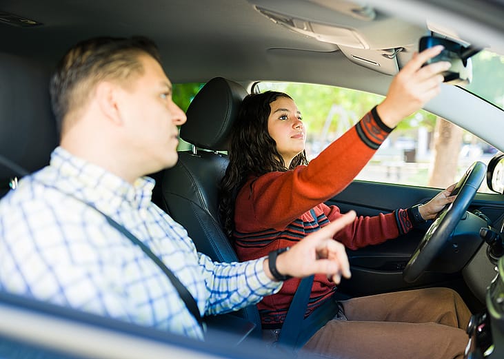 A teenage girl adjusting rearview mirror while learning to drive with her father.