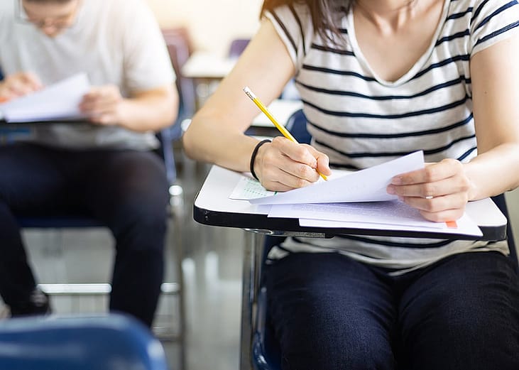 A female student sitting at a desk in a classroom while taking a test and writing on paper.