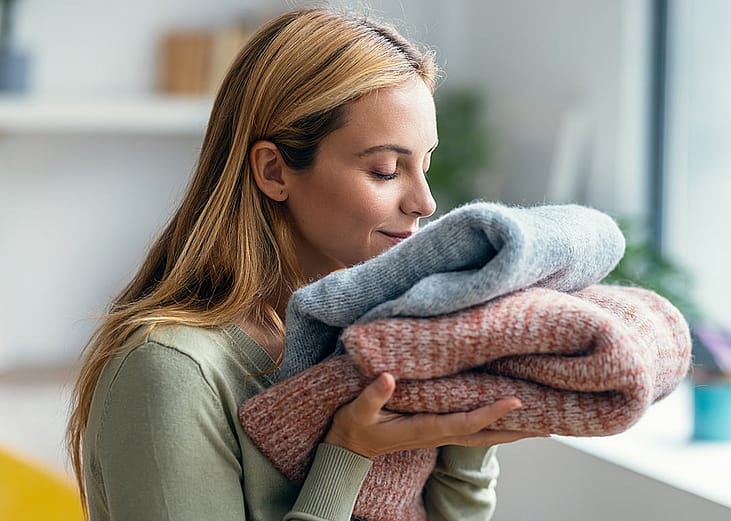 A beautiful young woman holding and smelling clean clothes at home.