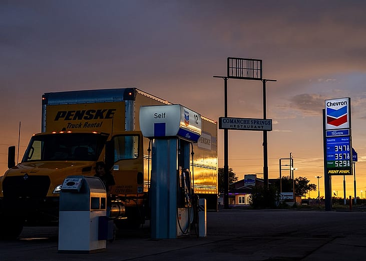 A truck getting fuel at a gasoline station with gas and diesel prices displayed at the back.