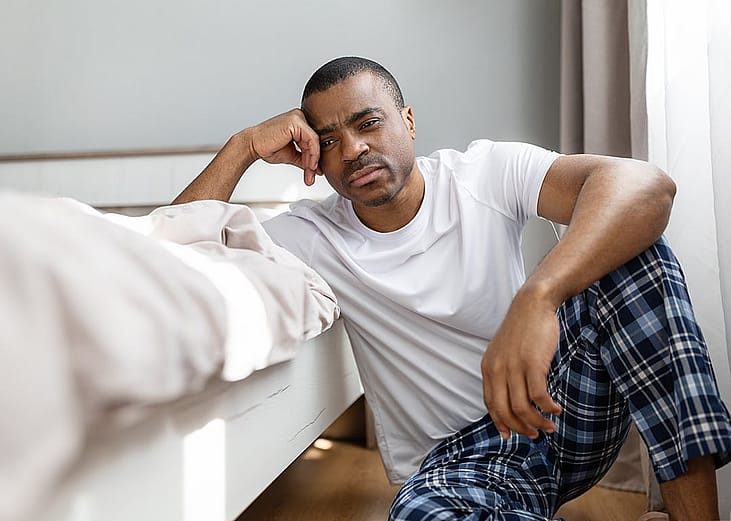 A man sitting by the end of his bed with a tired expression.