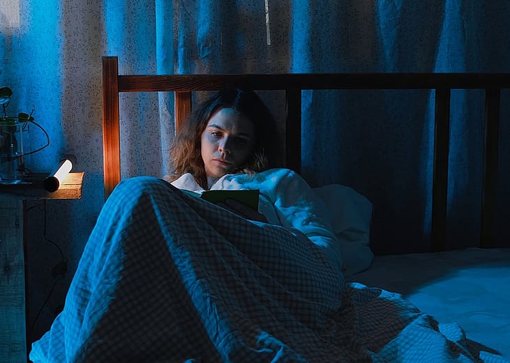 A young adult suffering from insomnia while reading a book in bed at night.