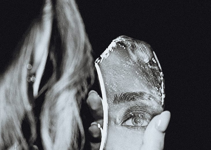 A black-and-white portrait of a woman holding a broken piece of a mirror that reflects a close-up of her eye.