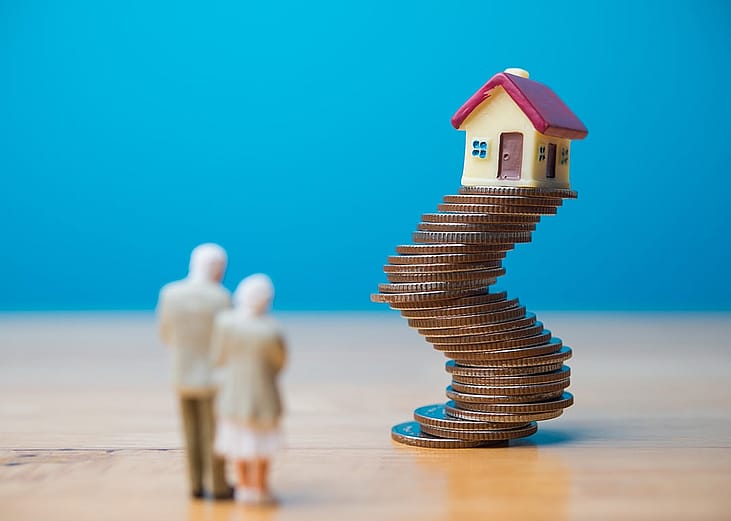 A miniature senior couple standing before a toy house on unstable stack of coins, representing financial risk with longer loans during retirement.