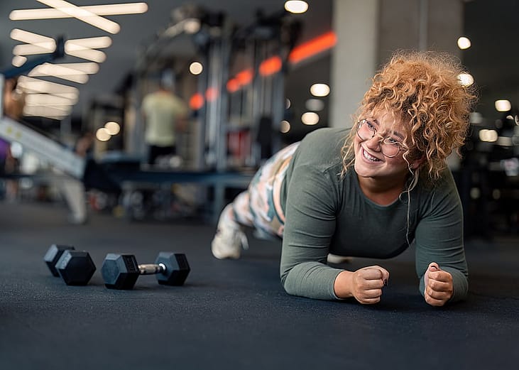 A young woman performing a forearm plank exercise on a gym floor.