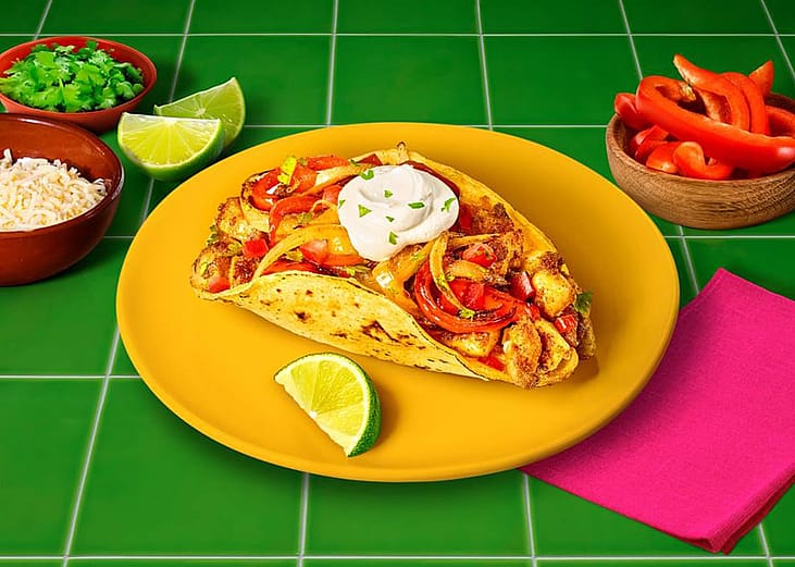 A fajita topped with sour cream served in a plate with lemon wedges, grated cheese, cilantro leaves, and bell peppers on the sides.