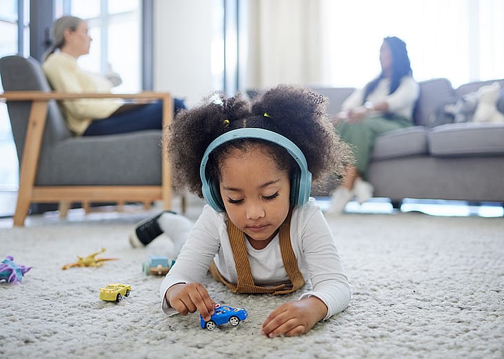 a young girl lying on a carpet and playing with toy cars with her parent in the background talking to a counselor.